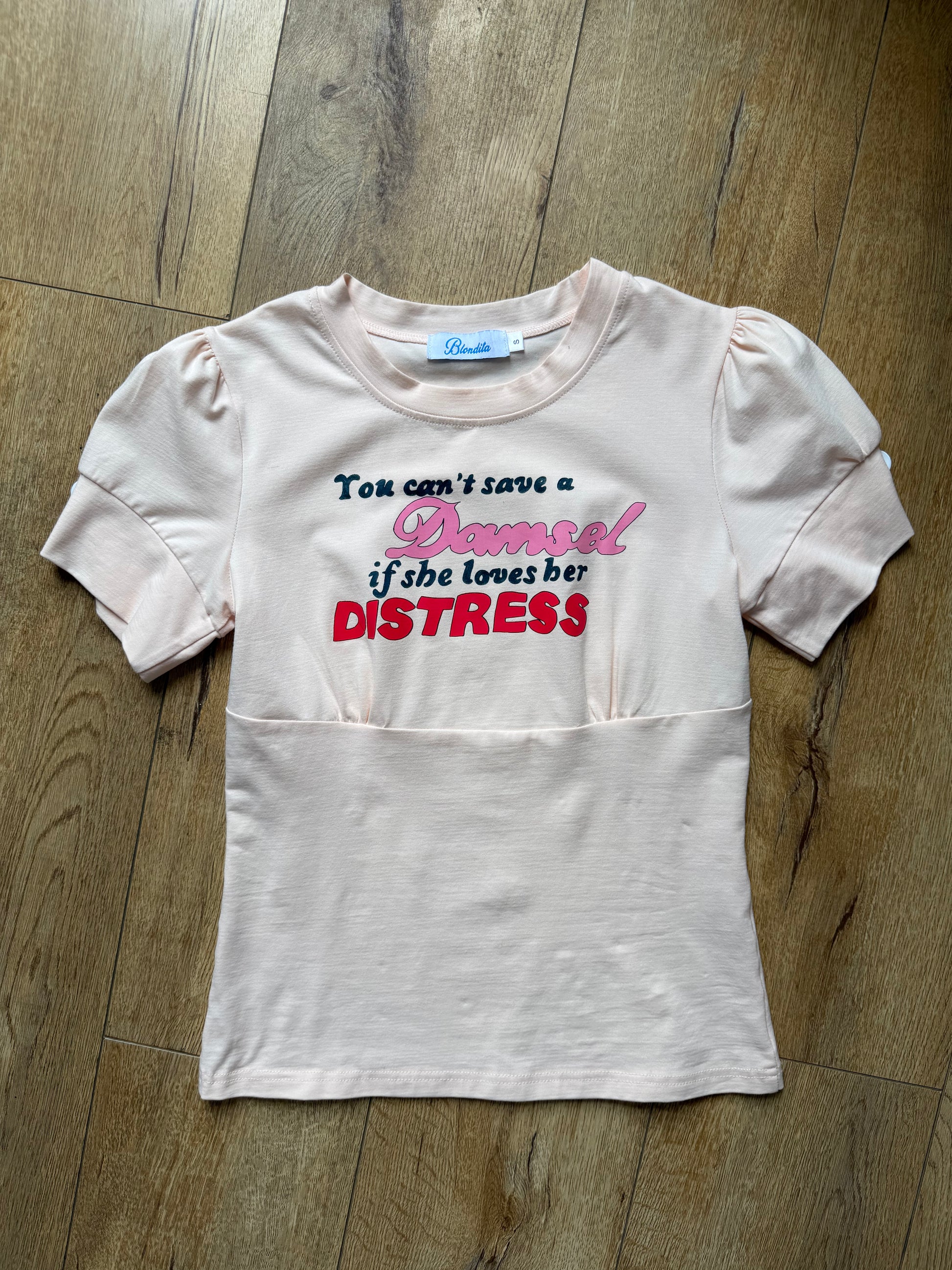 peach pink t shirt with buttons on the sleeve and a graphic that says "you can’t save a damsel if she loves her distress"